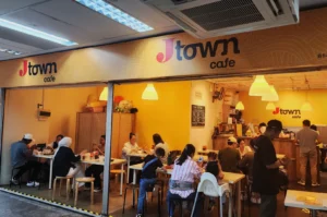 Bustling café scene with patrons seated at tables enjoying their meals. The interior is warmly lit with yellow walls and hanging lamps, creating a cozy ambiance.