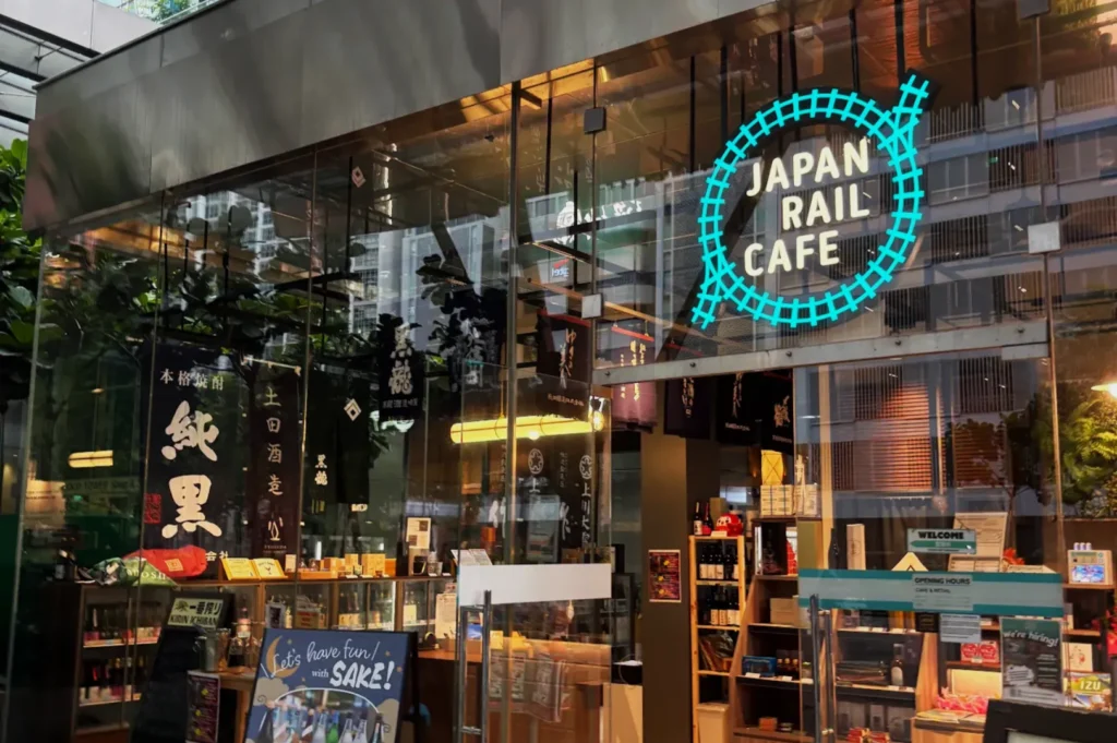 Exterior of Japan Rail Cafe, featuring a bright neon sign with a blue train track design. Glass facade showcases shelves and a “Have fun with Sake!” board.