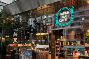 Exterior of Japan Rail Cafe, featuring a bright neon sign with a blue train track design. Glass facade showcases shelves and a “Have fun with Sake!” board.