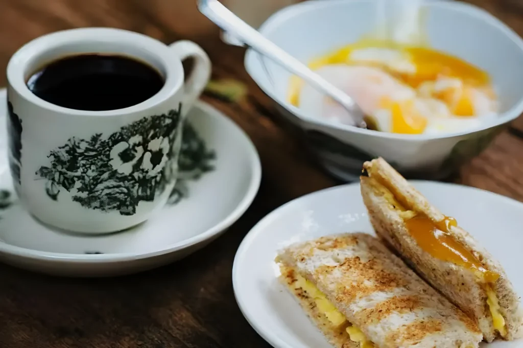 A cozy breakfast scene with a cup of black coffee in a floral cup, a bowl of soft-boiled eggs, and a plate of toasted bread with melted cheese.