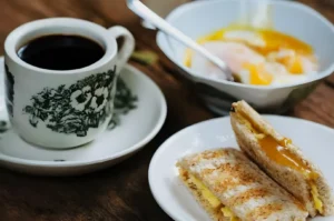 A cozy breakfast scene with a cup of black coffee in a floral cup, a bowl of soft-boiled eggs, and a plate of toasted bread with melted cheese.