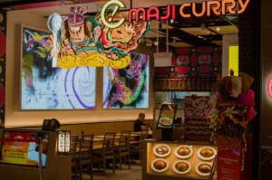 Maji Curry restaurant entrance with vibrant anime art on walls, illuminated curry dish models, seating area, and a colorful flower bouquet on the right.