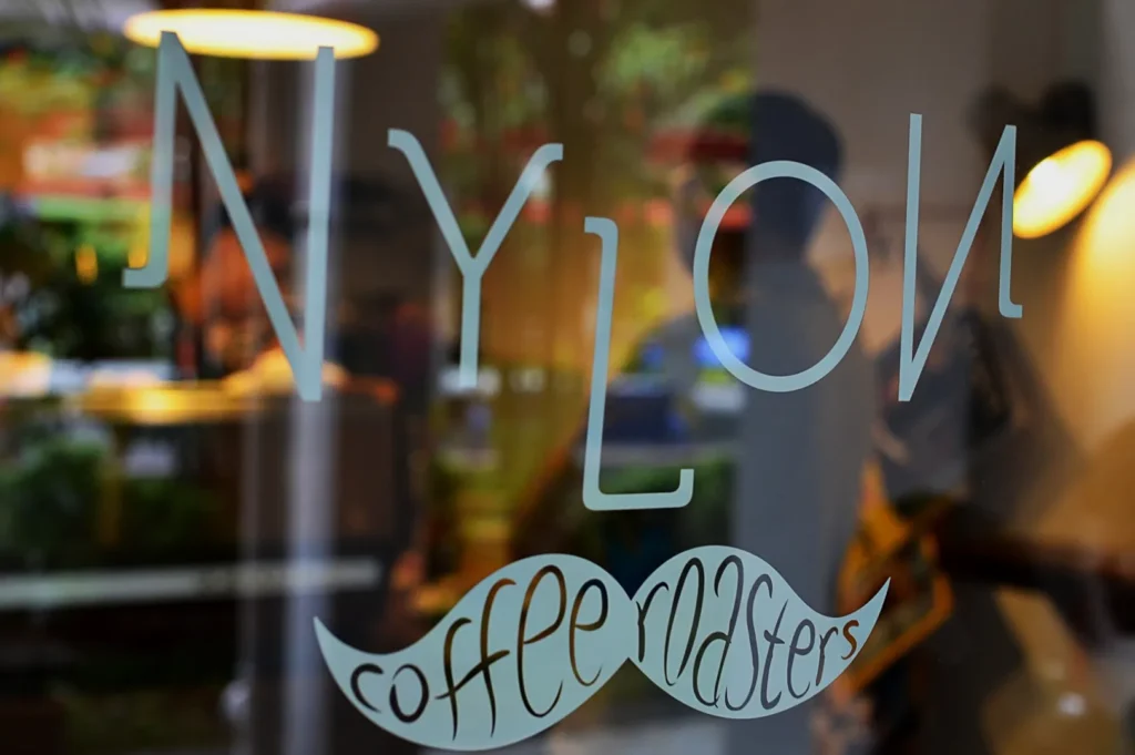 Glass door with "NYLON" in large letters and "coffee roasters" in a mustache shape below. Warm indoor lighting and blurred greenery outside.