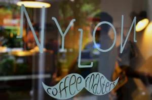 Glass door with "NYLON" in large letters and "coffee roasters" in a mustache shape below. Warm indoor lighting and blurred greenery outside.