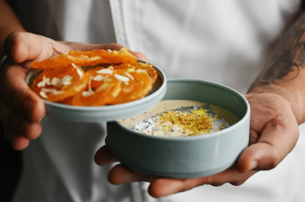 Hands holding a light blue dish with jalebi topped with almonds and a matching bowl of creamy dessert garnished with silver leaf and pistachios.