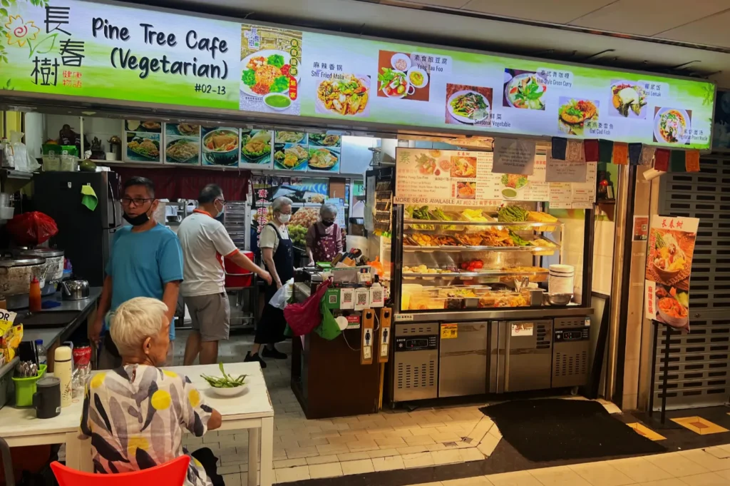 Vegetarian cafe with bright signage displaying food images. Inside, staff prepare dishes. A patron sits at a table, creating a welcoming vibe.