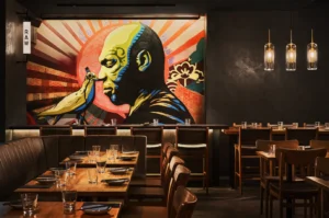 A modern restaurant with wooden tables and chairs, featuring a striking mural of a person's profile against vibrant red rays. The atmosphere is warm and artistic.