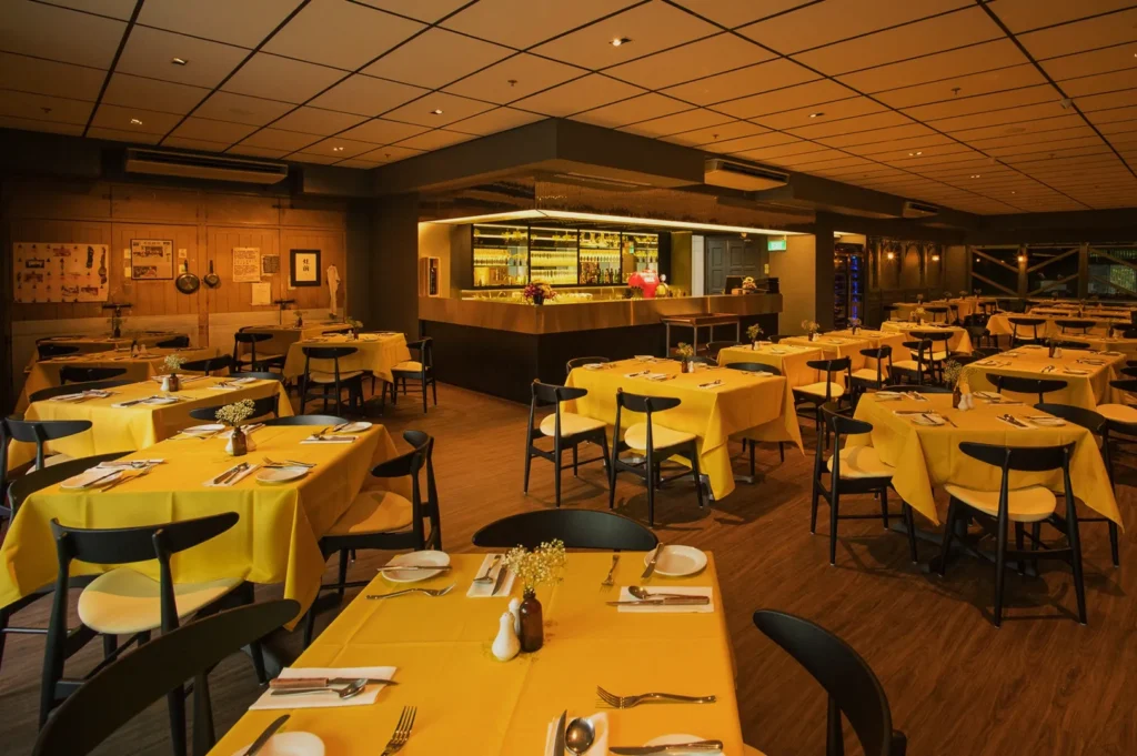 Cozy restaurant with yellow tablecloths, black chairs, and neatly set tables. Warm lighting and wooden decor create an inviting, relaxed ambiance.