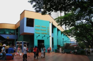 A lively scene at Tekka Centre in Singapore, showcasing a colorful building with people walking on a tiled plaza, surrounded by lush trees and greenery.