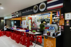 Casual Thai eatery with red stools and long tables, and a counter displaying menu photos. Customers dining; staff in green prepares food. Cozy ambiance.