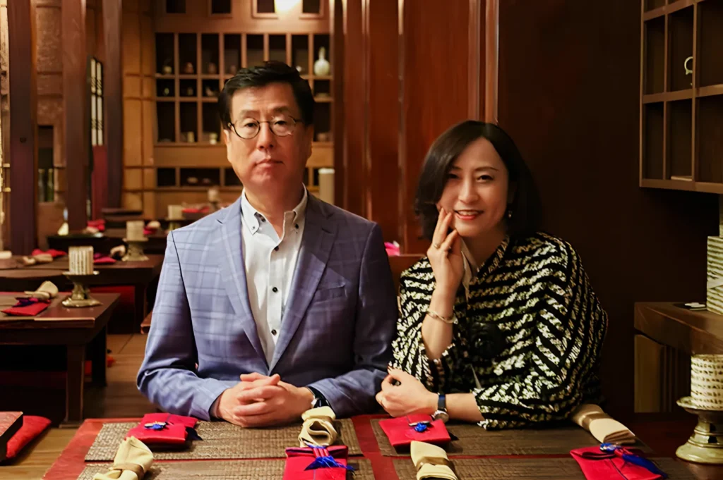 A couple sits at a restaurant table, elegantly set with red napkins and decorative items. The man wears a light blue suit, and the woman wears a patterned dress, smiling warmly. The ambiance is cozy with wooden decor, creating a sophisticated atmosphere.