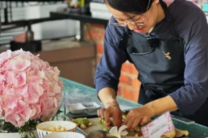 A person wearing glasses and a dark apron is focused on preparing food at a table. A large bouquet of pink flowers decorates the scene.