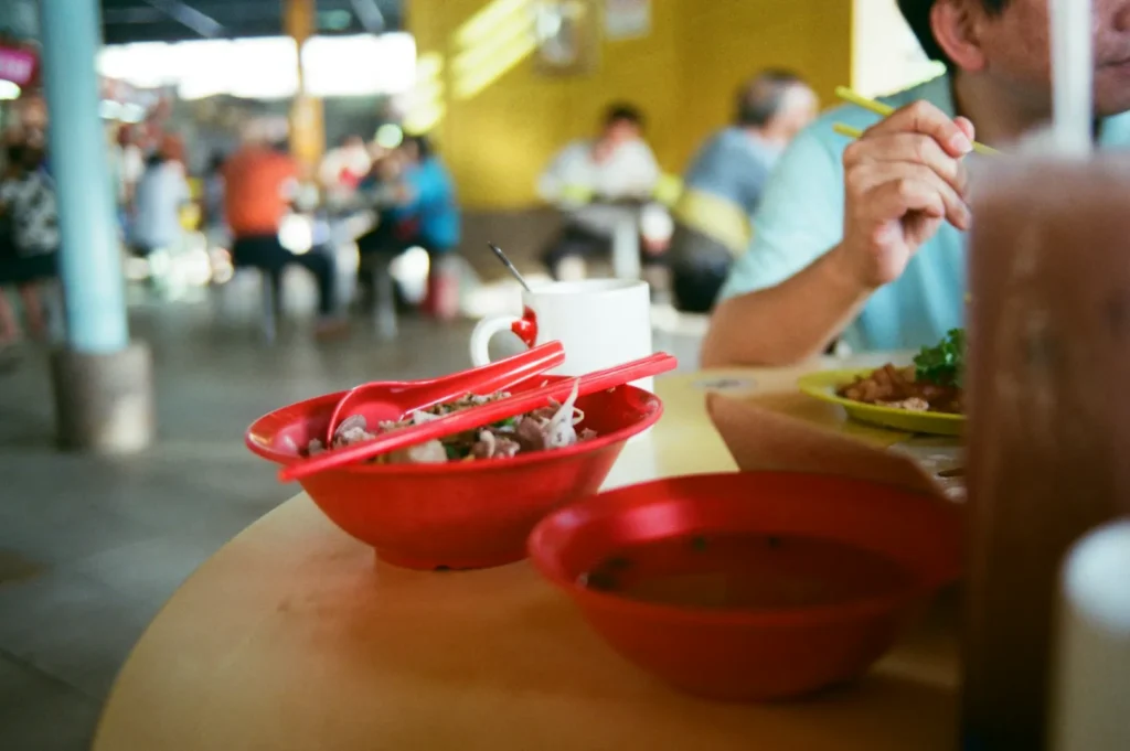 A colorful dining scene shows red bowls of noodles with chopsticks, a white mug, and a person eating. Background has people seated, creating a lively ambiance.