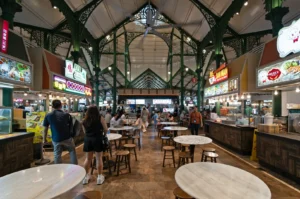 Interior of a lively food market with high ceilings and green iron arches. Various food stalls line the sides. People are walking and seated at round tables.