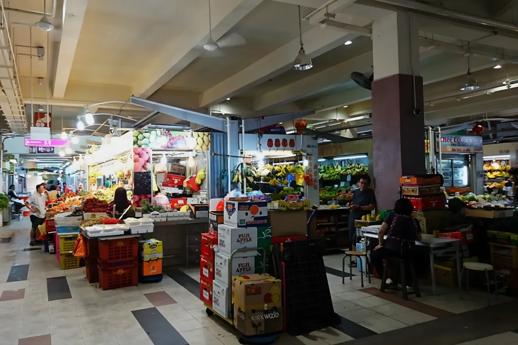 Indoor market scene with vibrant fruit stalls, stacked boxes, and shoppers browsing under warm lighting. A lively, bustling atmosphere prevails.