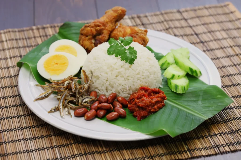 Plate with Nasi Lemak: rice topped with cilantro, boiled eggs, fried chicken, sliced cucumber, anchovies, peanuts, and sambal on banana leaf.