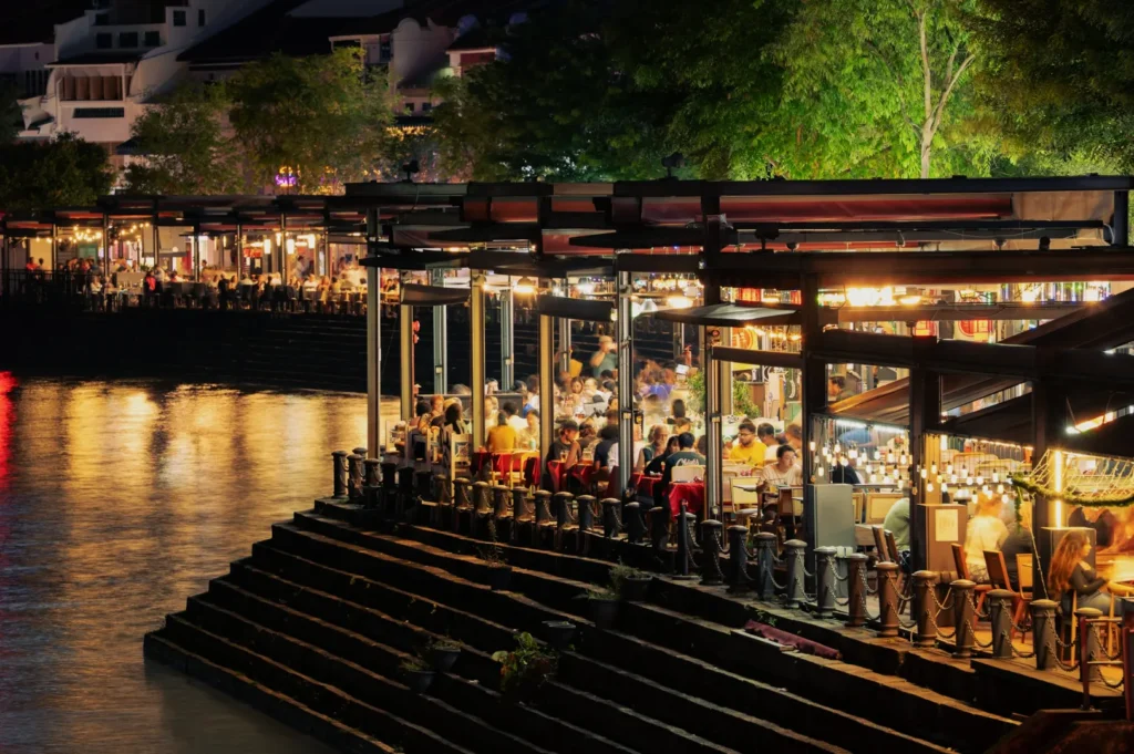 A lively riverside restaurant at night, with warmly lit dining areas filled with people enjoying meals. Reflections shimmer on the calm water nearby.