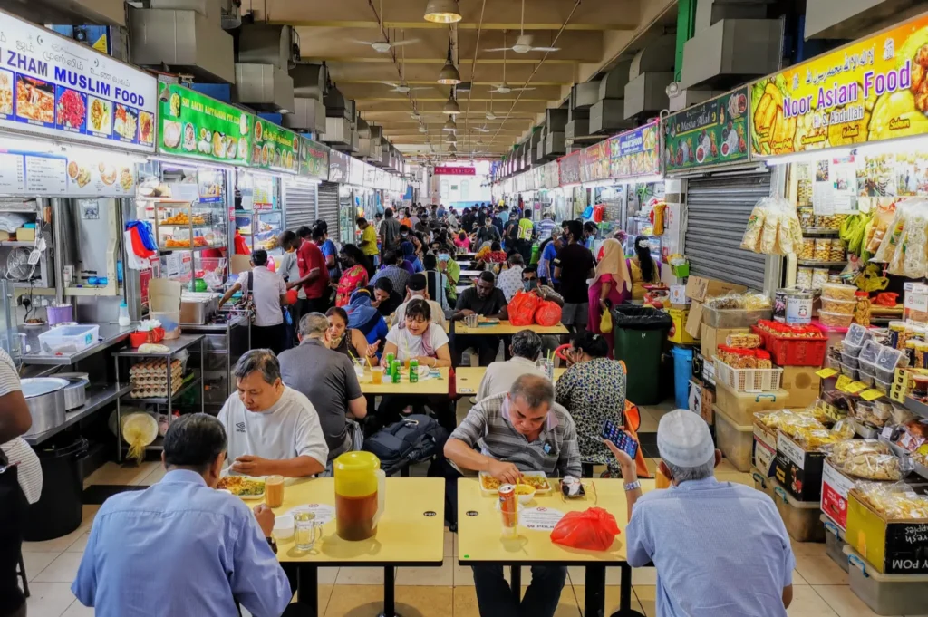 A busy indoor food market with diverse patrons at tables enjoying meals. Stalls line both sides, offering a vibrant array of Asian and Muslim cuisines.