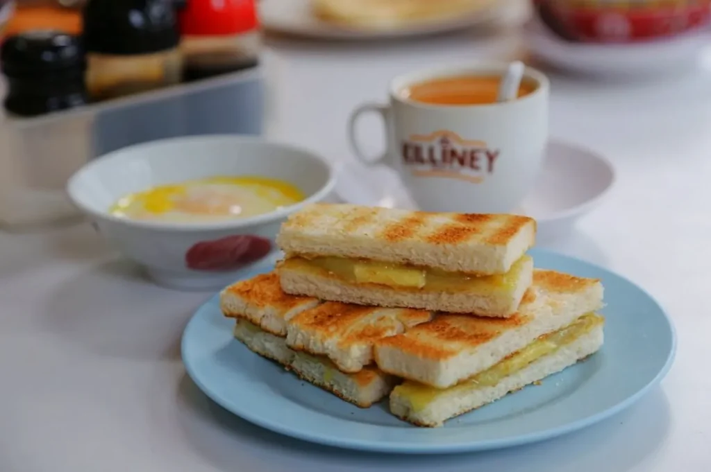 Toasted bread with butter and kaya on a blue plate, paired with a saucer of soft-boiled egg and a cup of coffee, creating a cozy breakfast setting.
