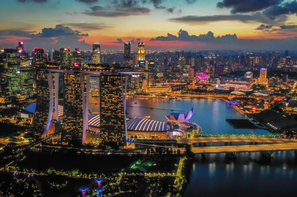Aerial view of Singapore at dusk, showcasing the illuminated Marina Bay Sands hotel, city skyline, and calm bay. The sky features a vibrant sunset.