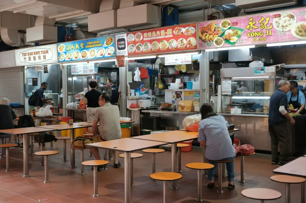 Hawker center interior with three open food stalls, each displaying colorful menus. People sit at orange stools around simple tables, creating a bustling, casual atmosphere.
