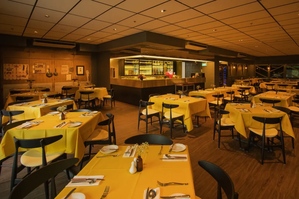 Spacious restaurant with soft lighting, yellow tablecloths, and neatly set tables. Bar in the background. Warm, inviting ambiance.