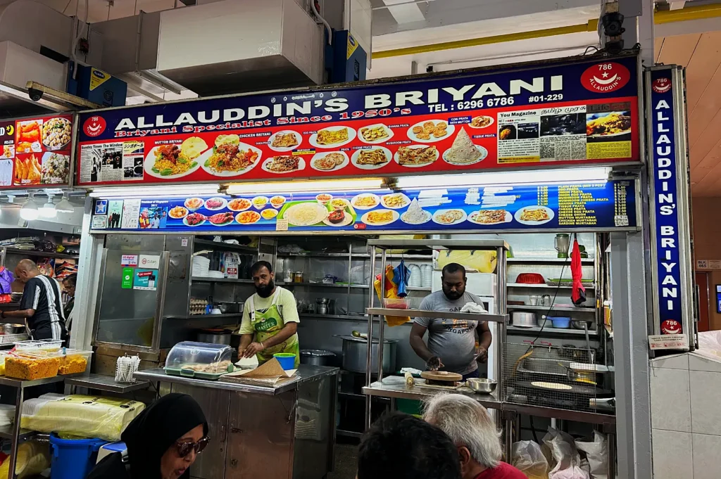 Stall at a food market named "Allauddin's Briyani" with vibrant menu images. Staff members are preparing food, creating a lively, aromatic atmosphere.