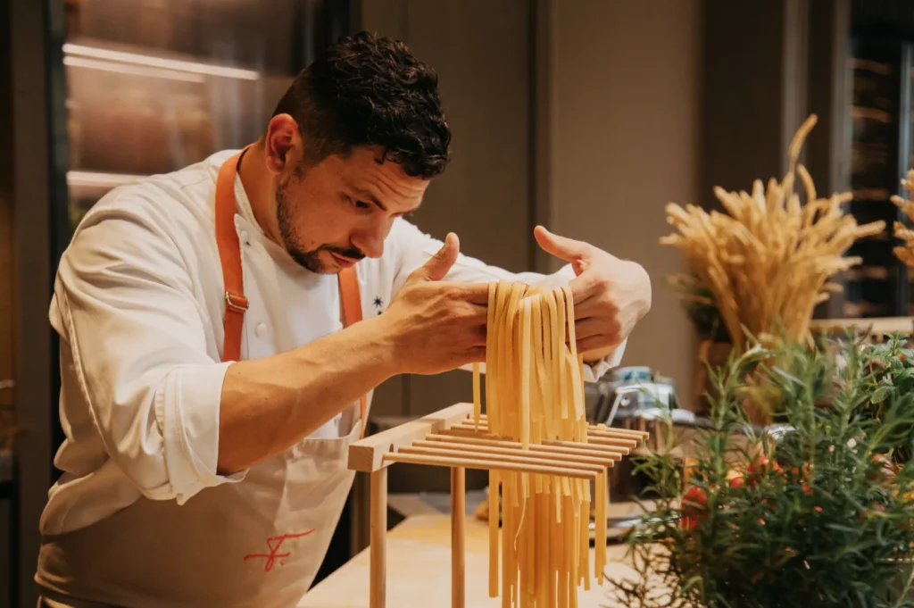 Chef in a white uniform carefully hangs fresh pasta on a wooden rack in a cozy kitchen, surrounded by herbs and wheat, focusing intently.