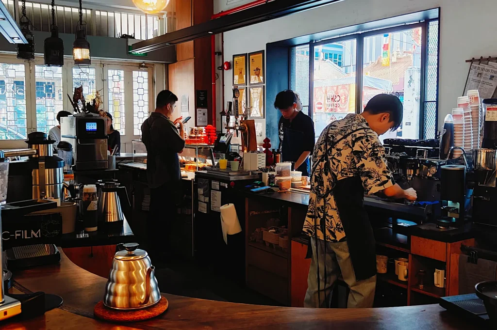 Cafe interior with a warm ambiance. Three baristas work behind the counter, making drinks. One uses an espresso machine, while two others prepare orders.