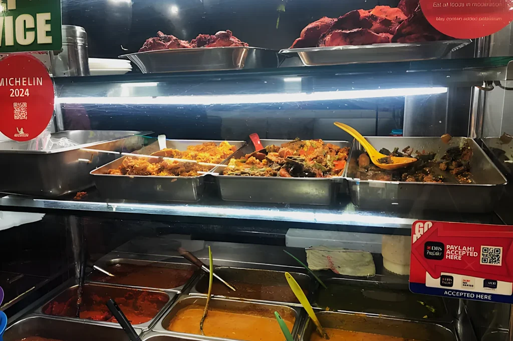 Buffet-style food display in a warm-lit setting with various colorful dishes in metal trays, marked by a Michelin 2024 plaque, conveying quality and variety.