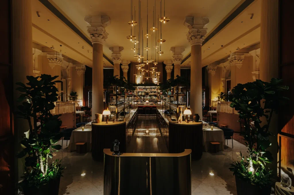 Elegant restaurant interior with grand columns, soft ambient lighting, and lush greenery. Rows of hanging pendant lights create a warm, inviting atmosphere.