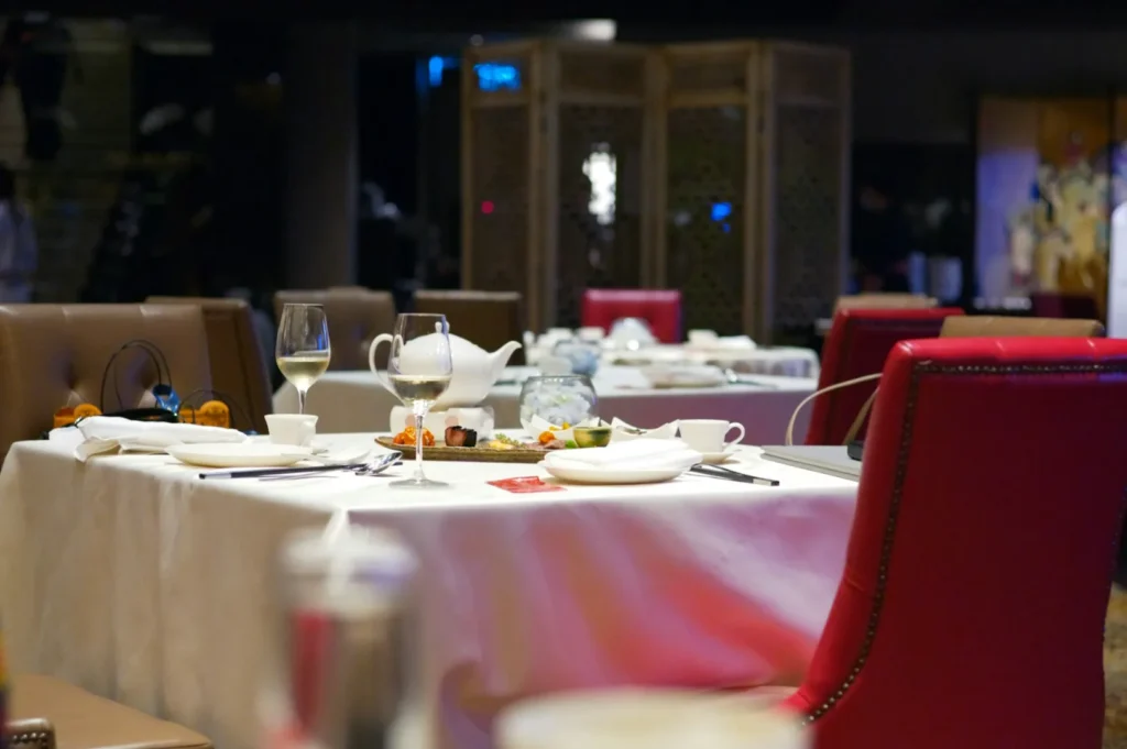 Elegant restaurant setting with tables adorned with white tablecloths, wine glasses, and plates. Red and brown chairs add a cozy, inviting ambiance.