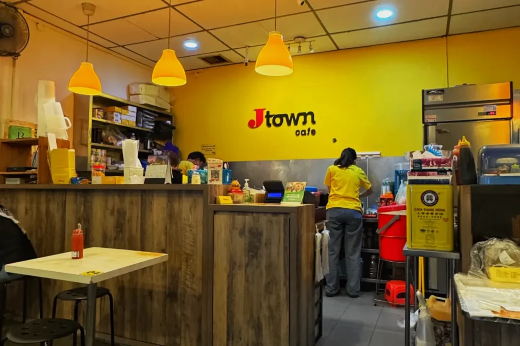 Cozy café interior with yellow walls, wooden counters, and hanging orange lights. Staff in casual attire work behind the counter, evoking a warm atmosphere.
