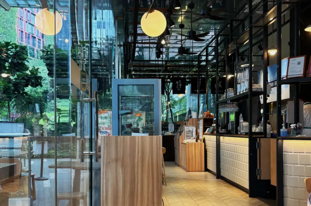 Modern cafe interior with wood details and white tiles. Large windows showcase lush outdoor greenery. Warm lighting creates a cozy atmosphere.