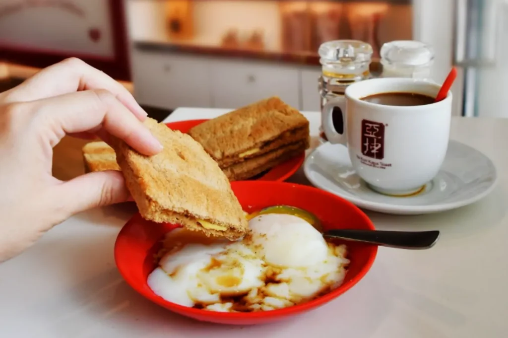 A hand dips kaya toast into soft-boiled eggs in a red bowl. Nearby, coffee in a white cup with a red spoon. The scene is warm and inviting.