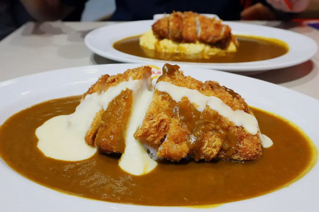 Close-up of two plates of Japanese curry featuring crispy fried cutlets on rice with creamy and curry sauces. The vibe is hearty and appetizing.