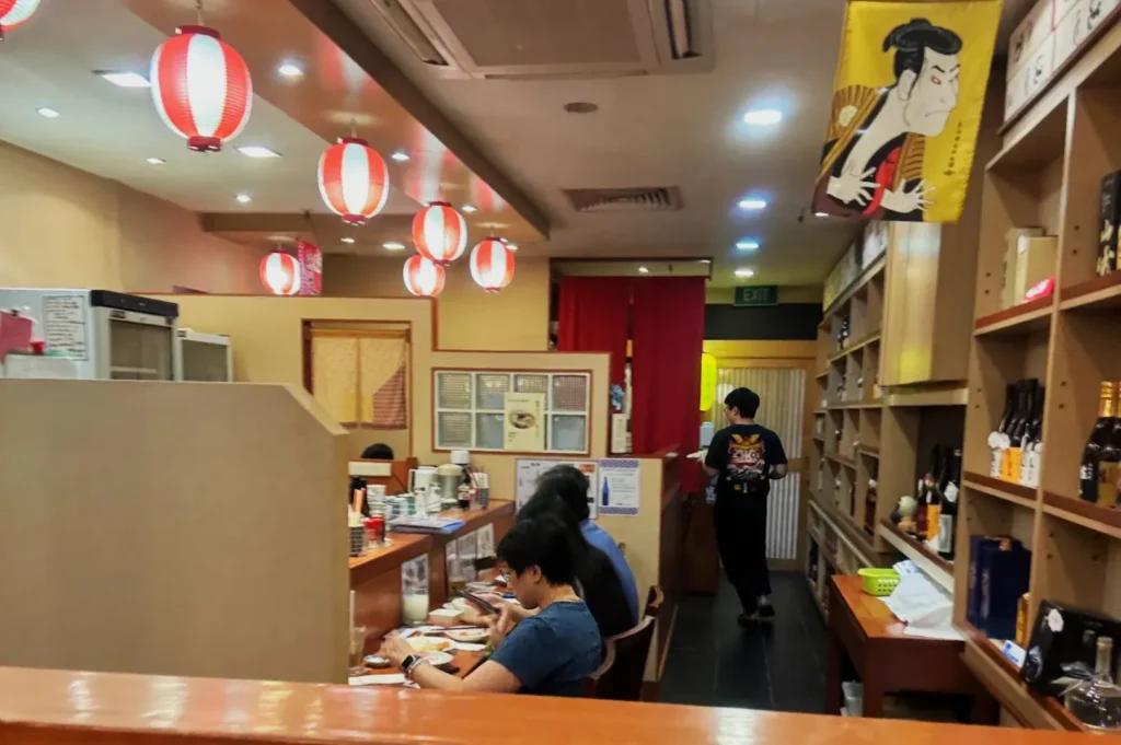 A cozy Japanese restaurant interior with red lanterns hanging from the ceiling. Customers sit at a counter on the left, while a staff member walks through an aisle. Shelves filled with bottles line the right wall, creating a warm and inviting atmosphere.