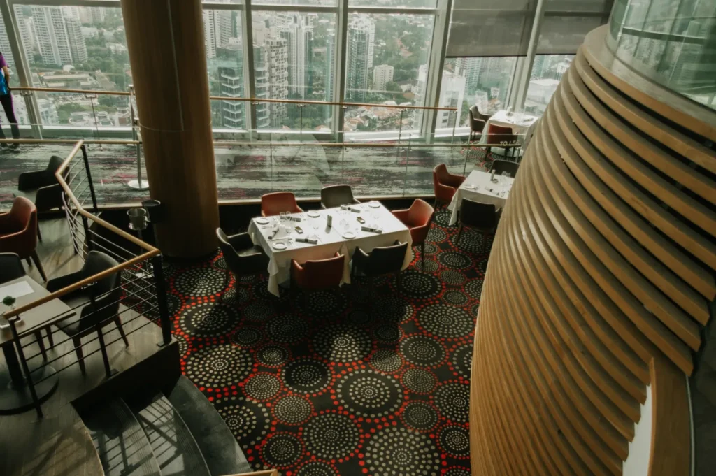 Modern, upscale restaurant interior with large windows overlooking a cityscape. Tables are set with white tablecloths, surrounded by patterned carpet.