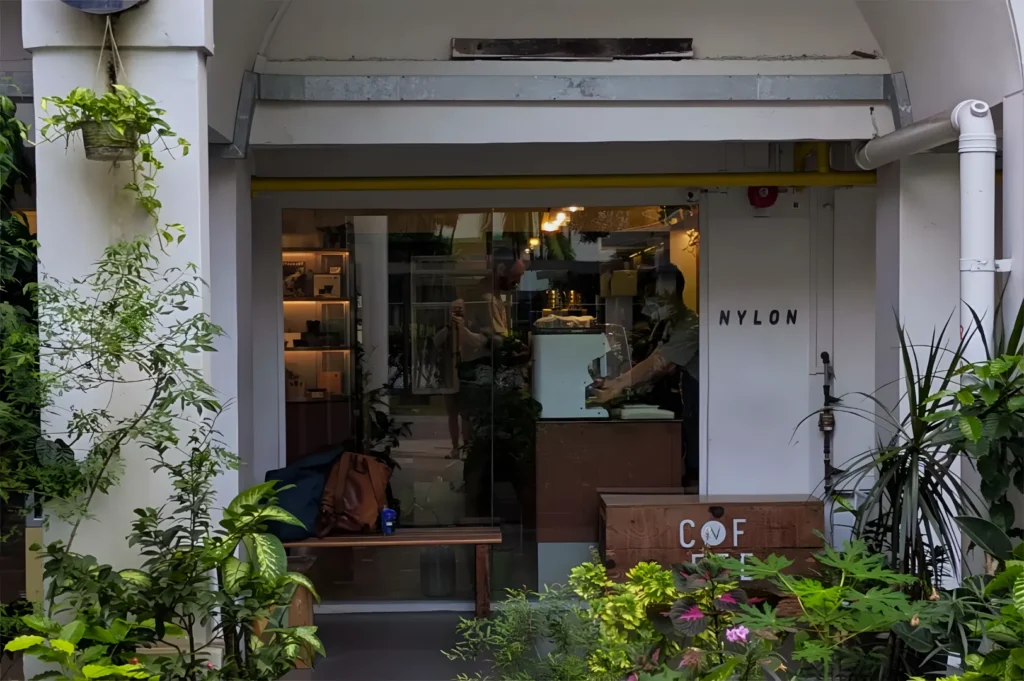 Street view of a cozy café with large windows displaying its modern interior. Lush green plants surround the entrance, creating a welcoming atmosphere.