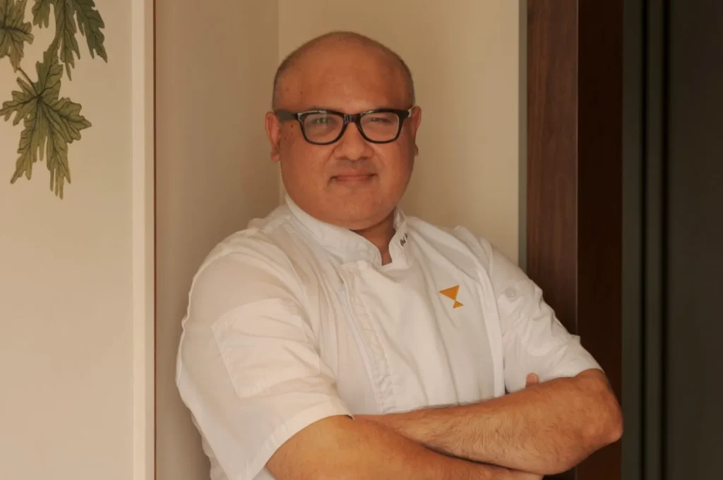 A chef in a white uniform stands confidently with arms crossed, wearing glasses. A calm expression, standing next to a wall with leafy decoration.