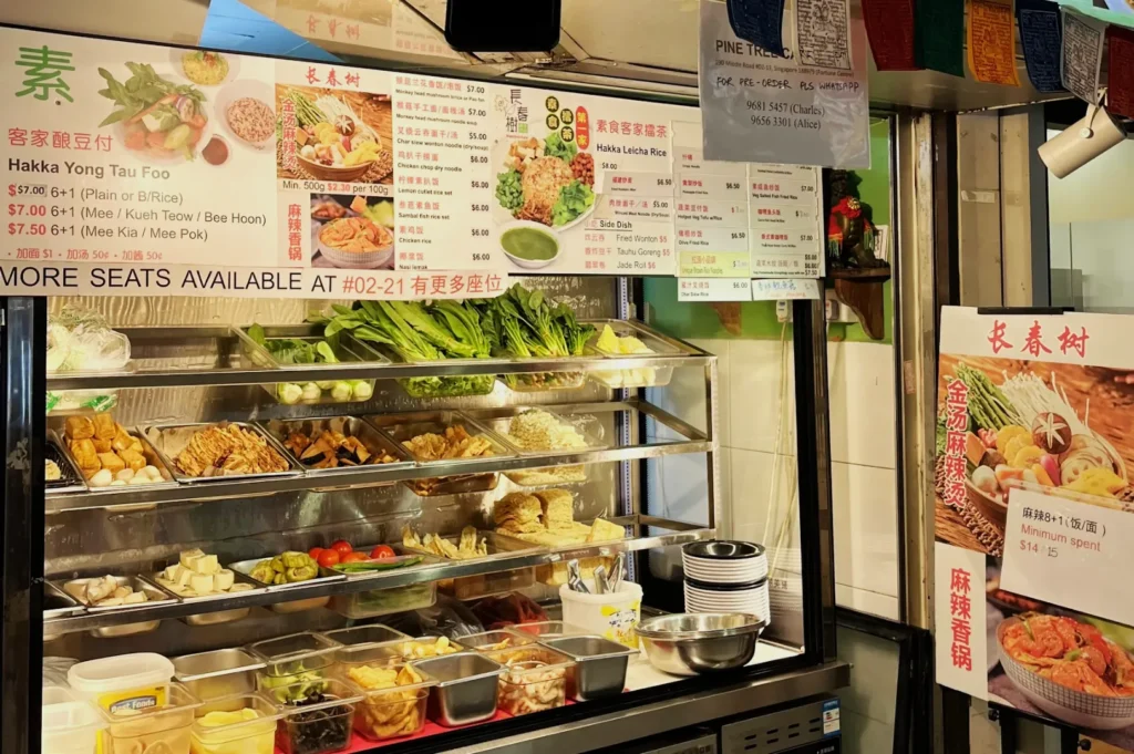 Food stall with a variety of ingredients displayed in metal trays, offering Hakka Yong Tau Foo. Colorful menus and signs with prices are visible above.
