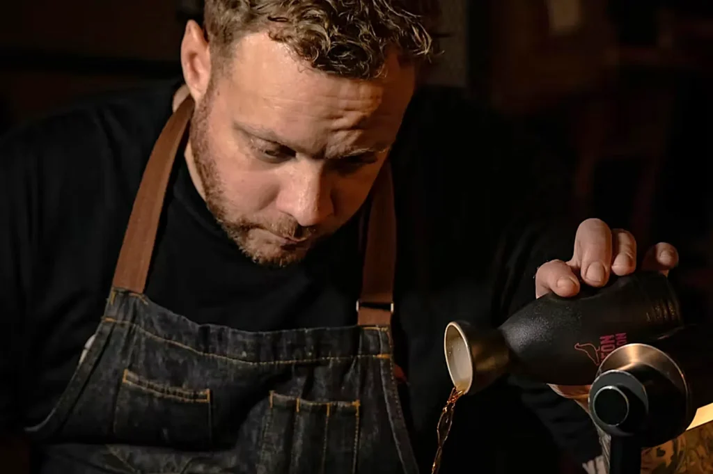 Chef Sean Mell in a dark apron pours liquid from a black vessel with focus and precision, creating a concentrated and thoughtful atmosphere.