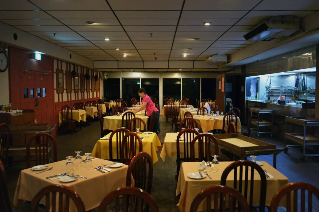 A person in a pink shirt sets tables in an empty restaurant with yellow tablecloths. The atmosphere is calm and organized, with dim lighting.