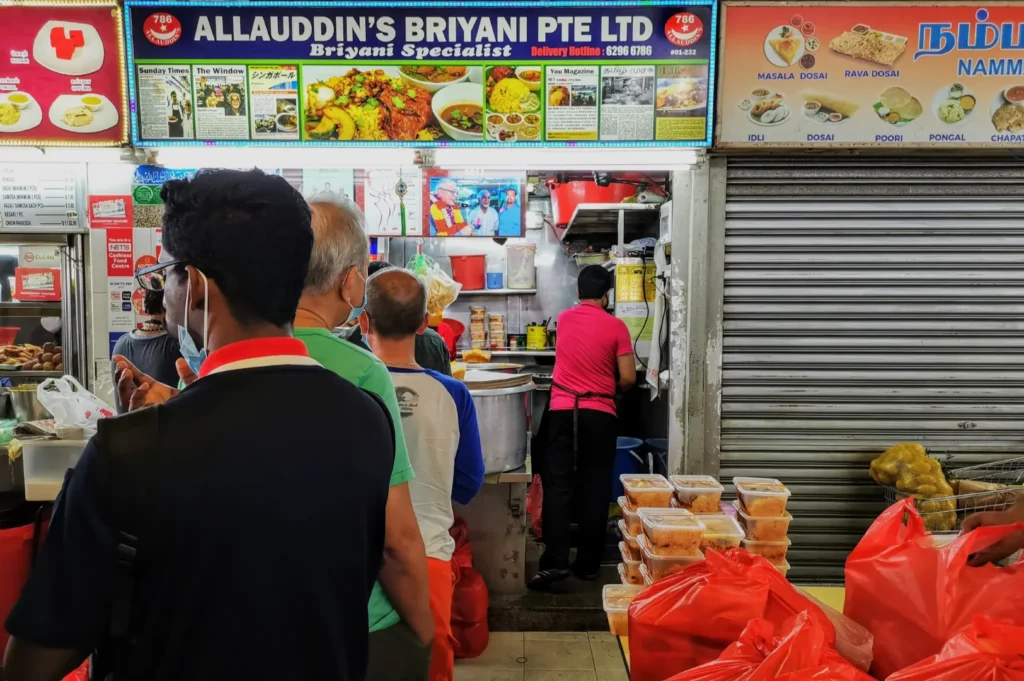 Line of people at a food stall named "Allauddin's Briyani," awaiting service. Tubs of food and red bags are stacked nearby, creating a busy atmosphere.