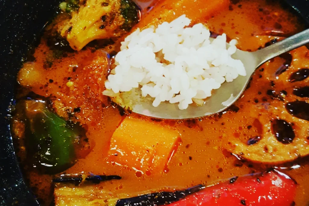 A bowl of vibrant vegetable curry with broccoli, carrots, and lotus root in rich broth. A spoon holds a portion of white rice, adding warmth.