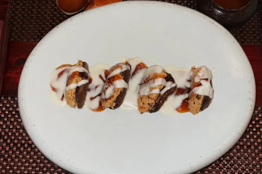 Five slices of stuffed dates drizzled with white and caramel sauces are arranged on an oval white plate, creating a sweet and elegant dessert display.