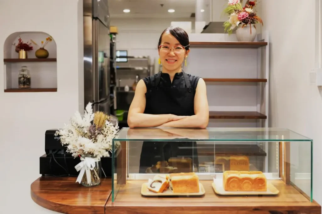 A smiling woman in a black apron stands behind a bakery counter with glass display, showcasing neatly arranged loaves of bread. Decor includes dried flowers, creating a welcoming atmosphere.