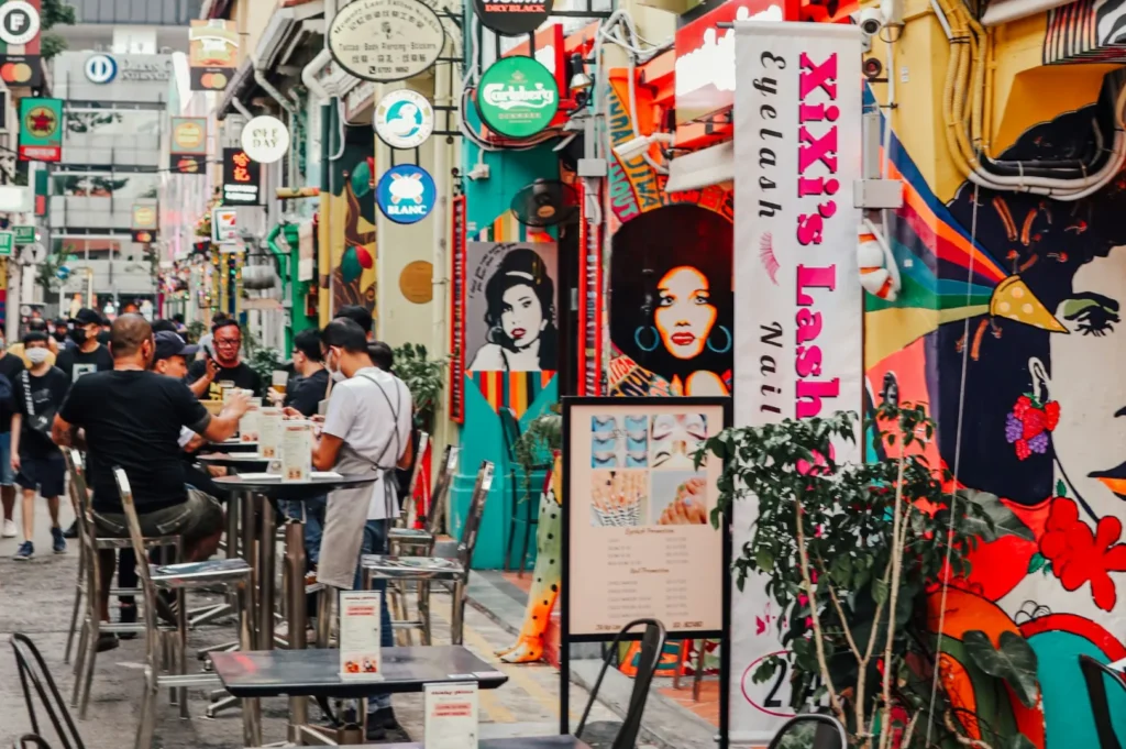 Vibrant street scene with people dining outdoors among colorful murals and bright signs. The lively atmosphere suggests a busy, cheerful urban setting.