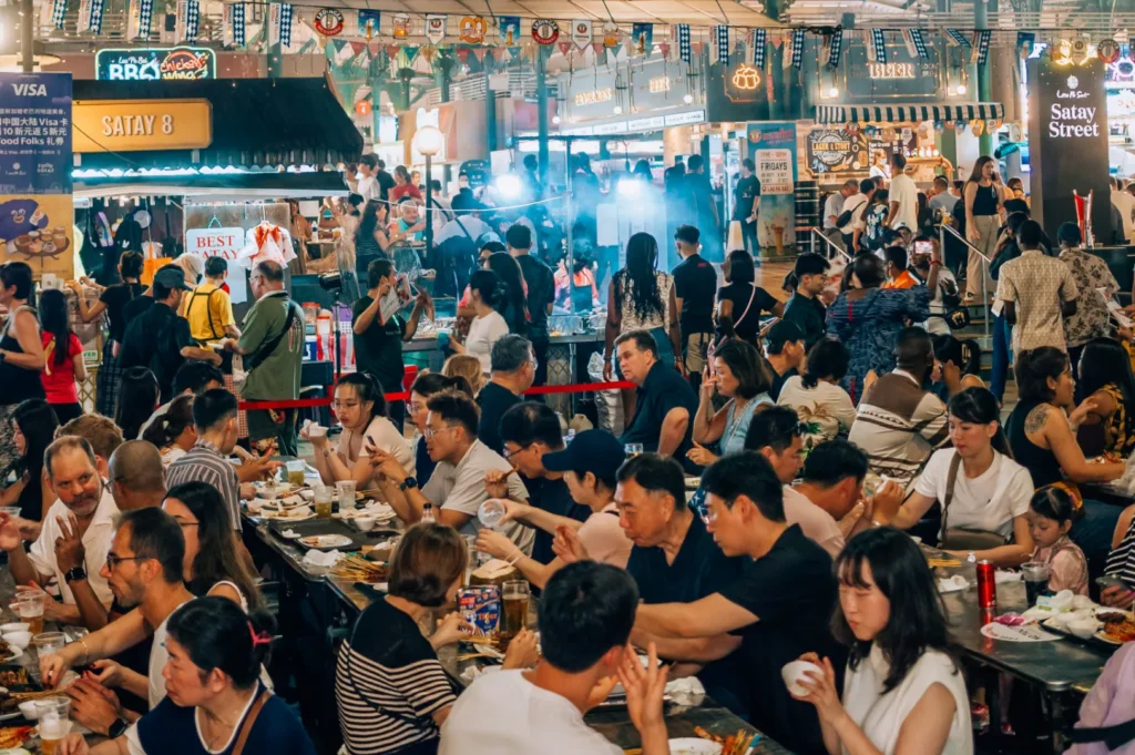 A bustling night market scene with diverse people enjoying food at shared tables. Vendors and bright signs line the background, creating a lively, festive atmosphere.