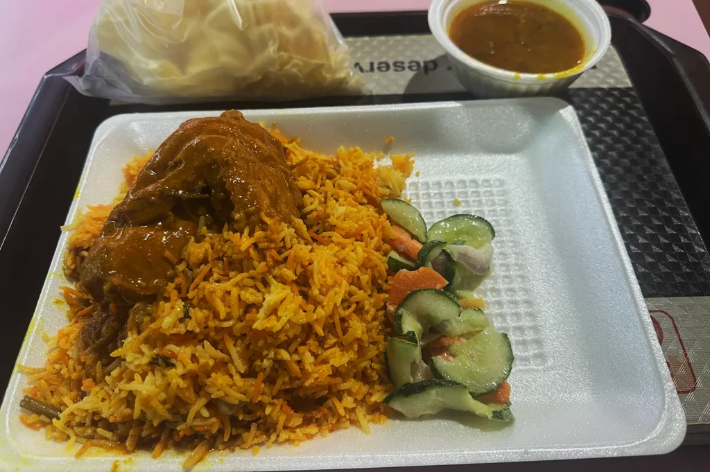 A white tray holds spicy chicken biryani with fragrant rice, fresh cucumber salad, and a bowl of curry sauce. Crispy papadum is wrapped in a bag.